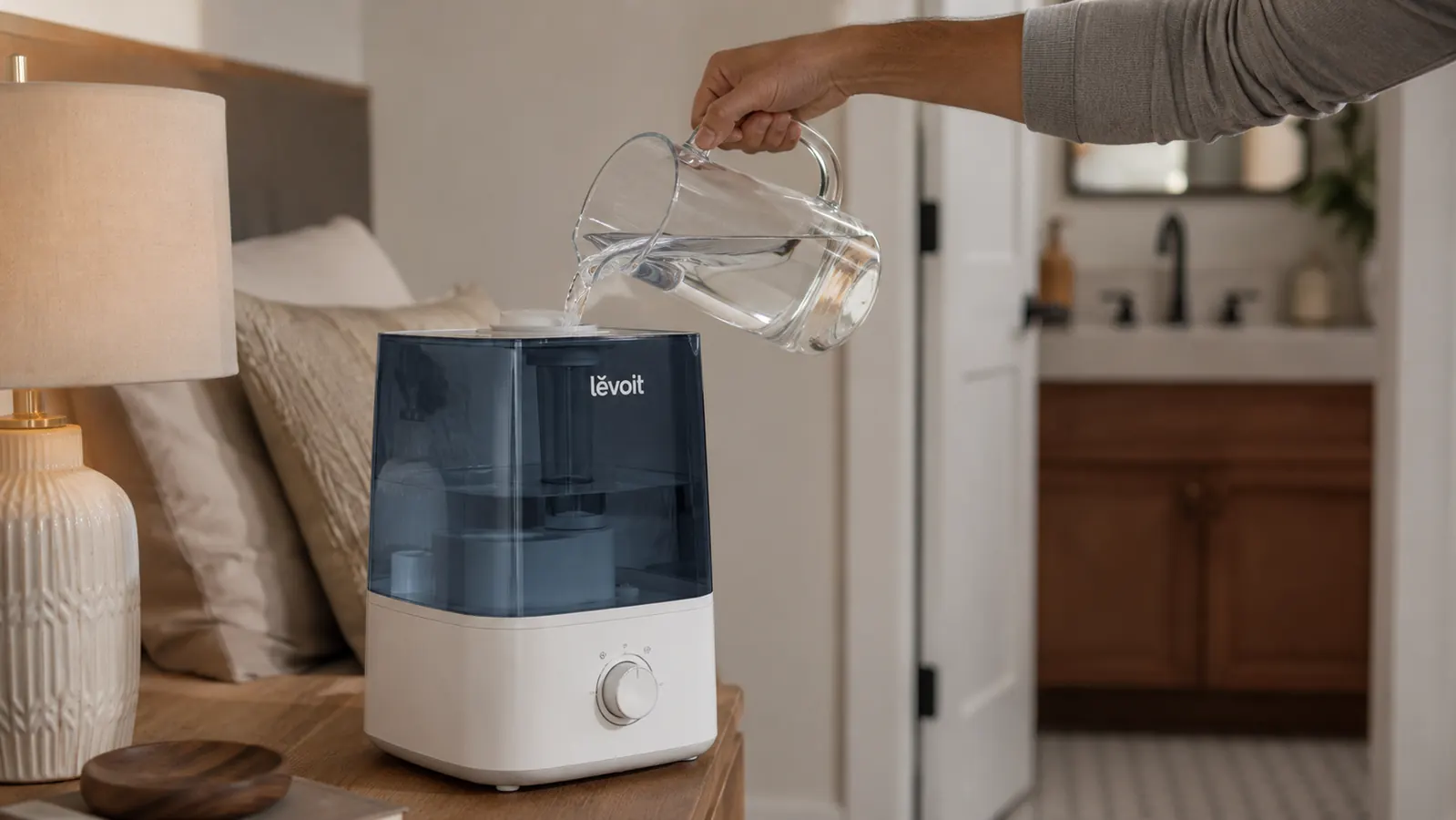 Hand tilting a cup of water into the top-fill opening of the Levoit humidifier tank at a kitchen sink
