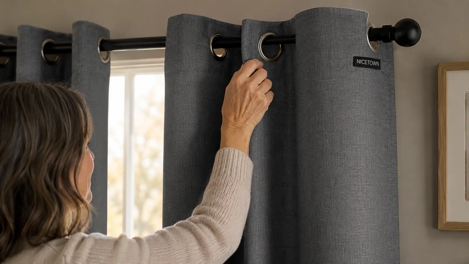 Hand closing a grey grommet blackout curtain along a black tension rod, using the actual NICETOWN panel as reference