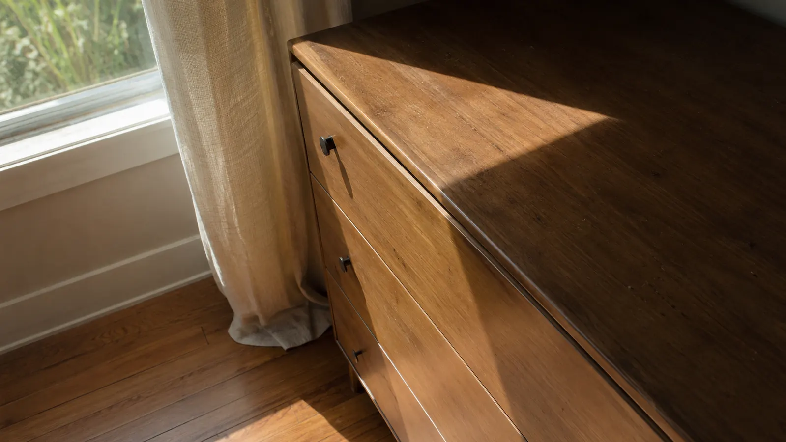 Sunlit dresser and wood floor near a window with the curtain partly open, showing an uneven patch of faded finish