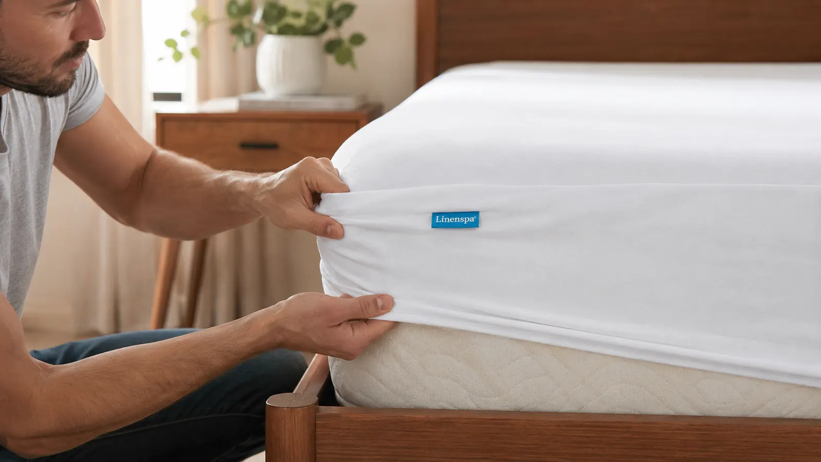 Hands stretching the elastic fitted corner of a white waterproof mattress protector down over a bare mattress corner