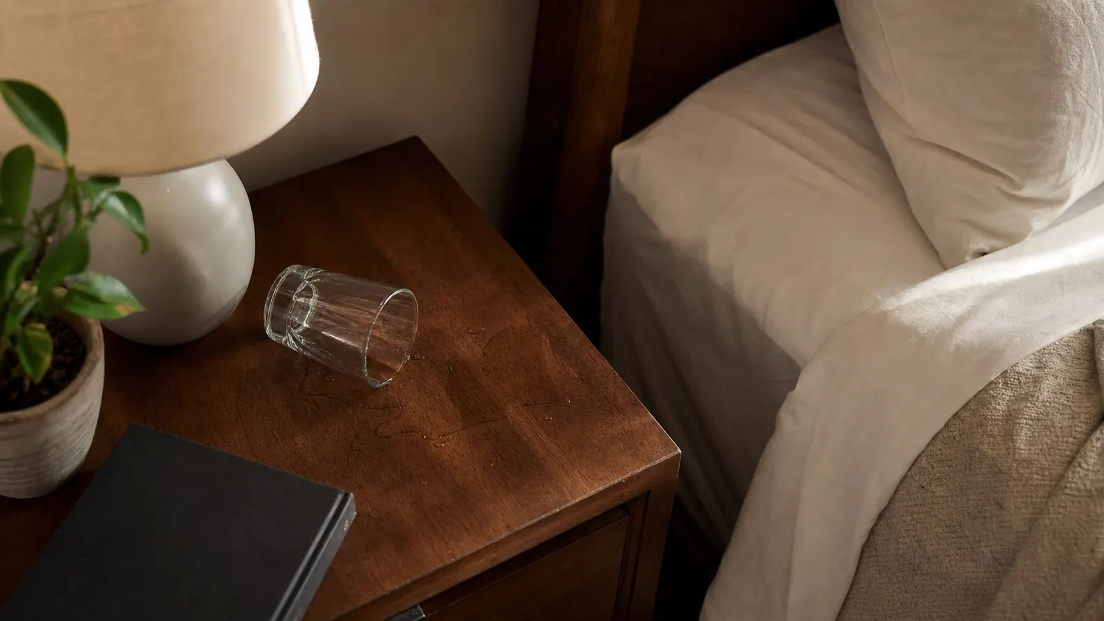 Overhead shot of a small water glass tipped over on a nightstand next to a made bed