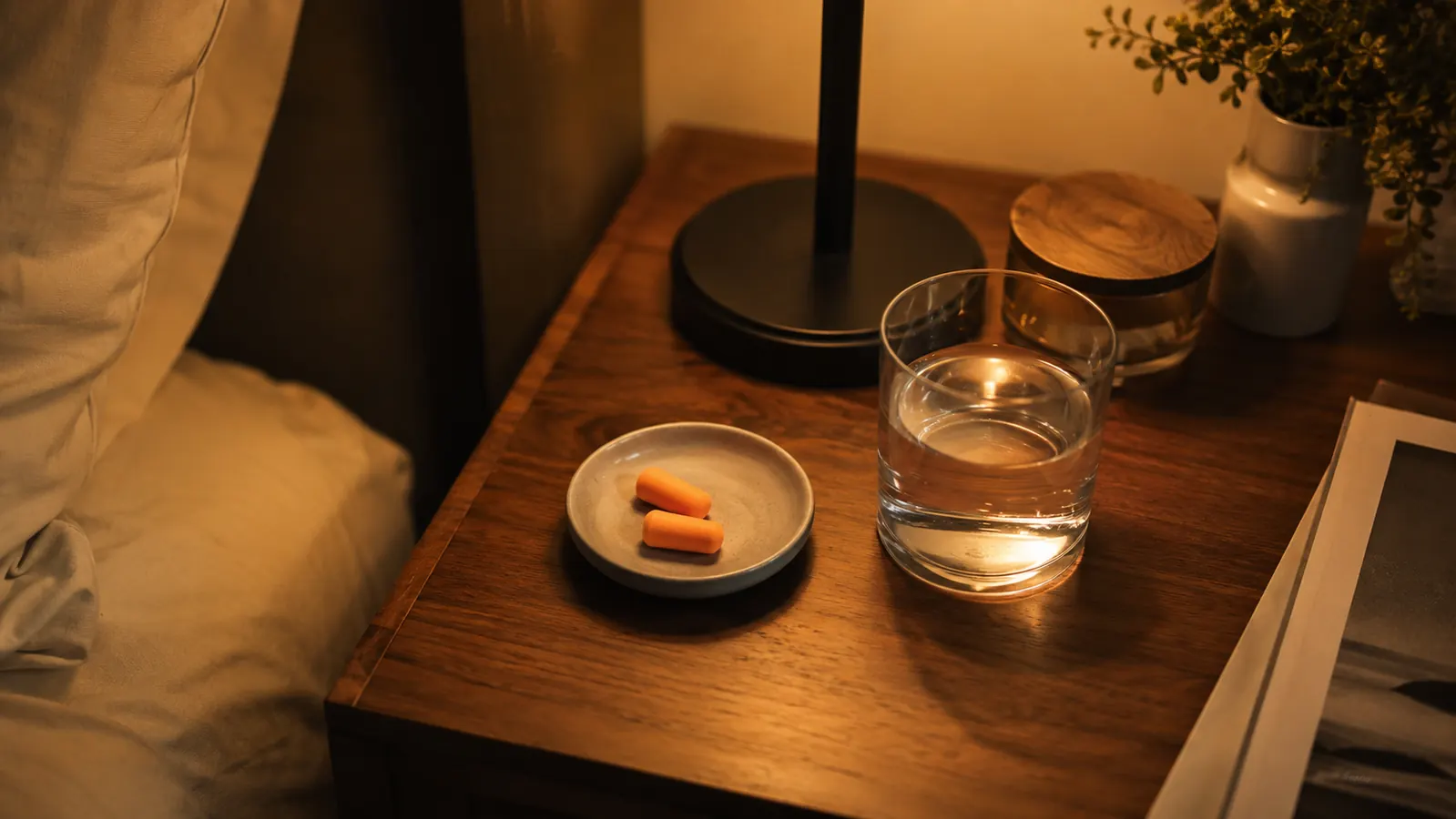 A small dish on a nightstand holding a few pairs of green foam ear plugs next to a lamp and a glass of water