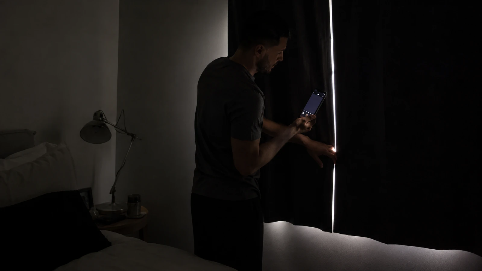 A person standing in a fully darkened bedroom at night holding up a phone with the camera flashlight off, checking the edges of a closed curtain for any visible glow