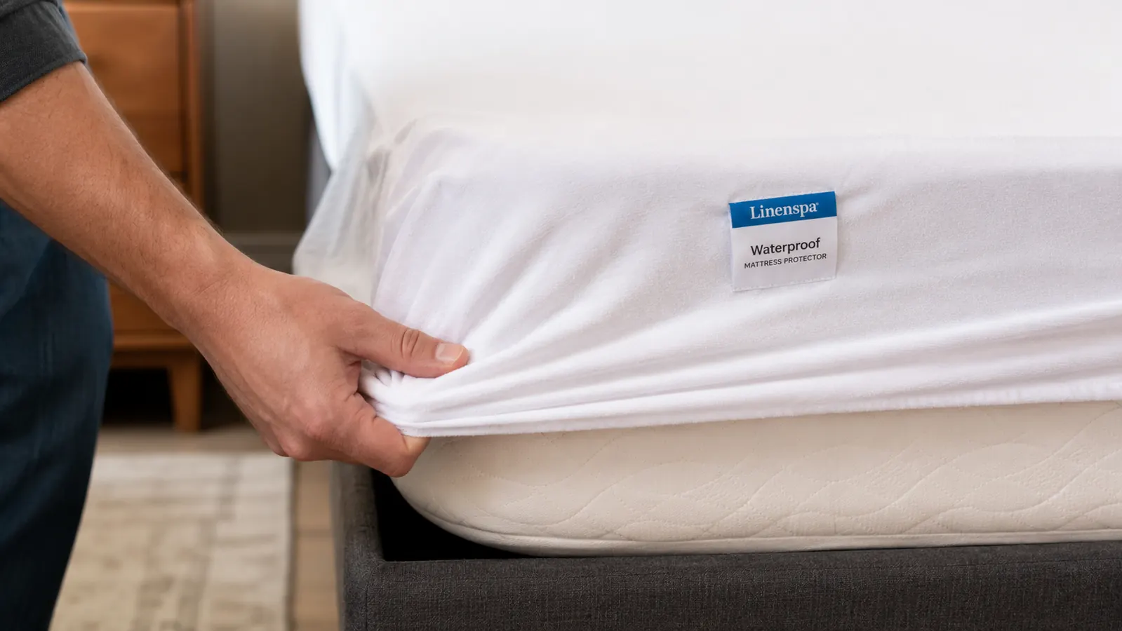A hand pulling a white fitted mattress protector over the corner of a bare mattress